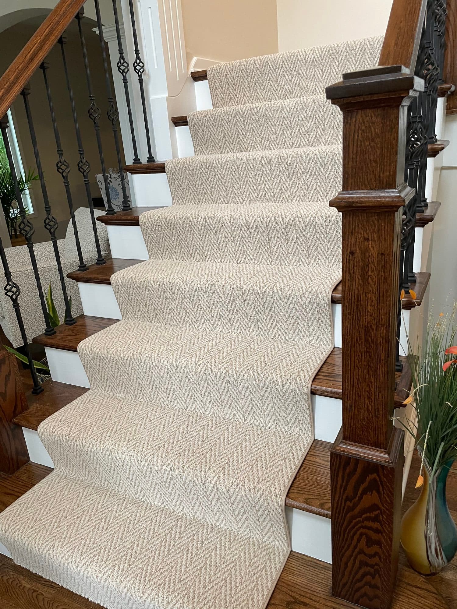 Cream and taupe herringbone carpet runner with bound edges on stained oak stairs with white risers and black iron balusters, seen from the bottom of the stairs beside the newel post